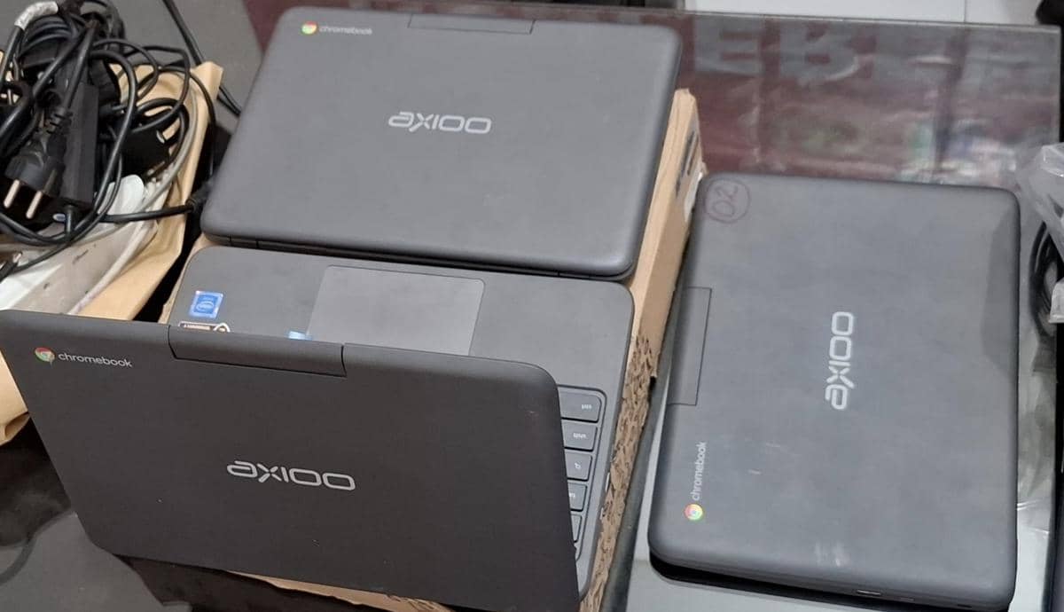 Beberapa laptop Chromebook merek Axioo berwarna hitam diletakkan di atas meja bersama kabel dan perlengkapan elektronik lainnya.