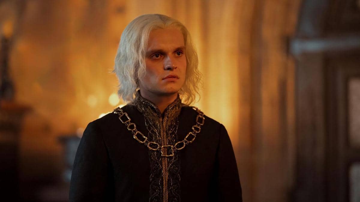 Seorang karakter berambut pirang mengenakan pakaian hitam berhias rantai emas dalam adegan serial House of the Dragon.