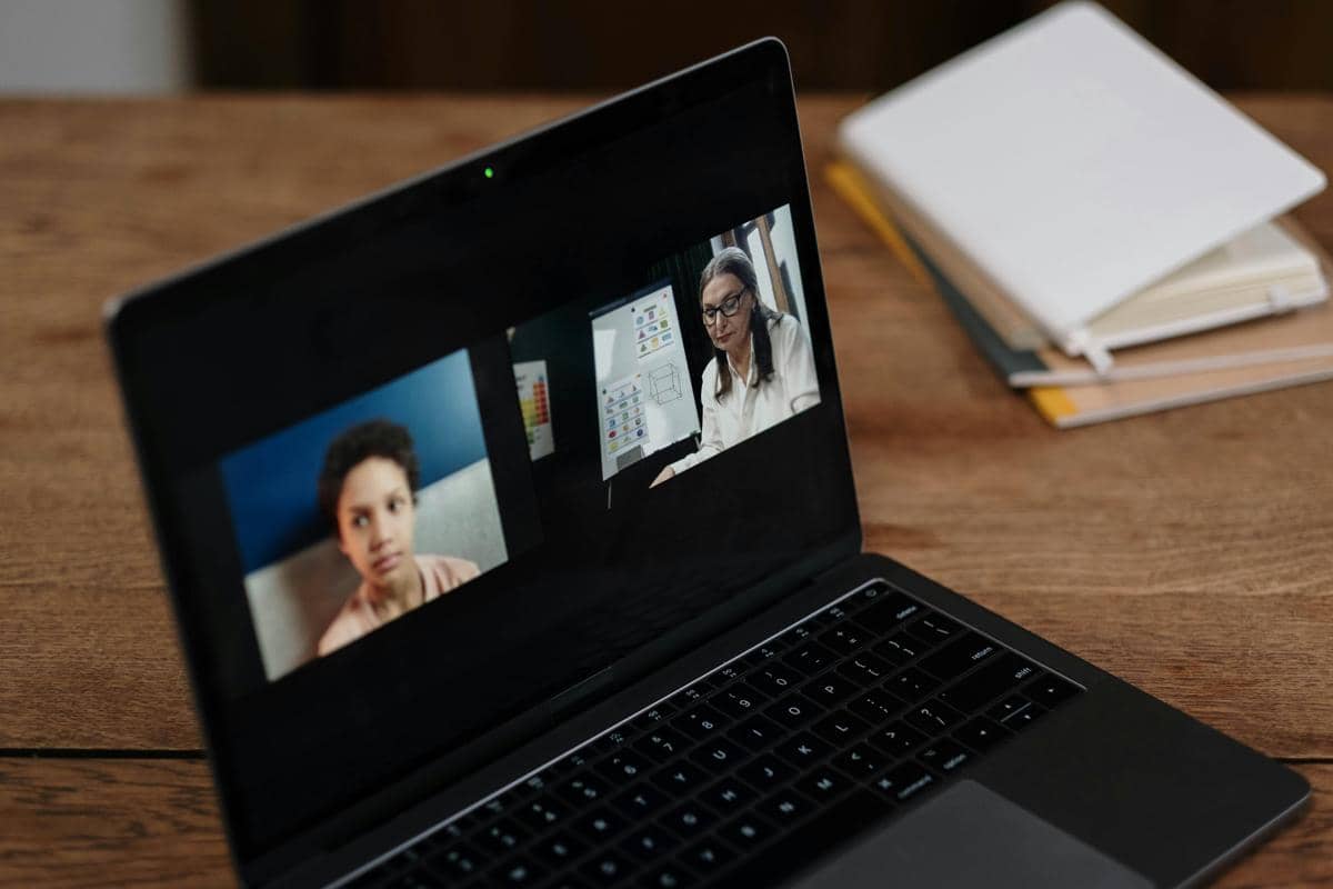 Layar laptop menampilkan dua orang sedang melakukan panggilan video, di atas meja kayu dengan beberapa buku di sampingnya.