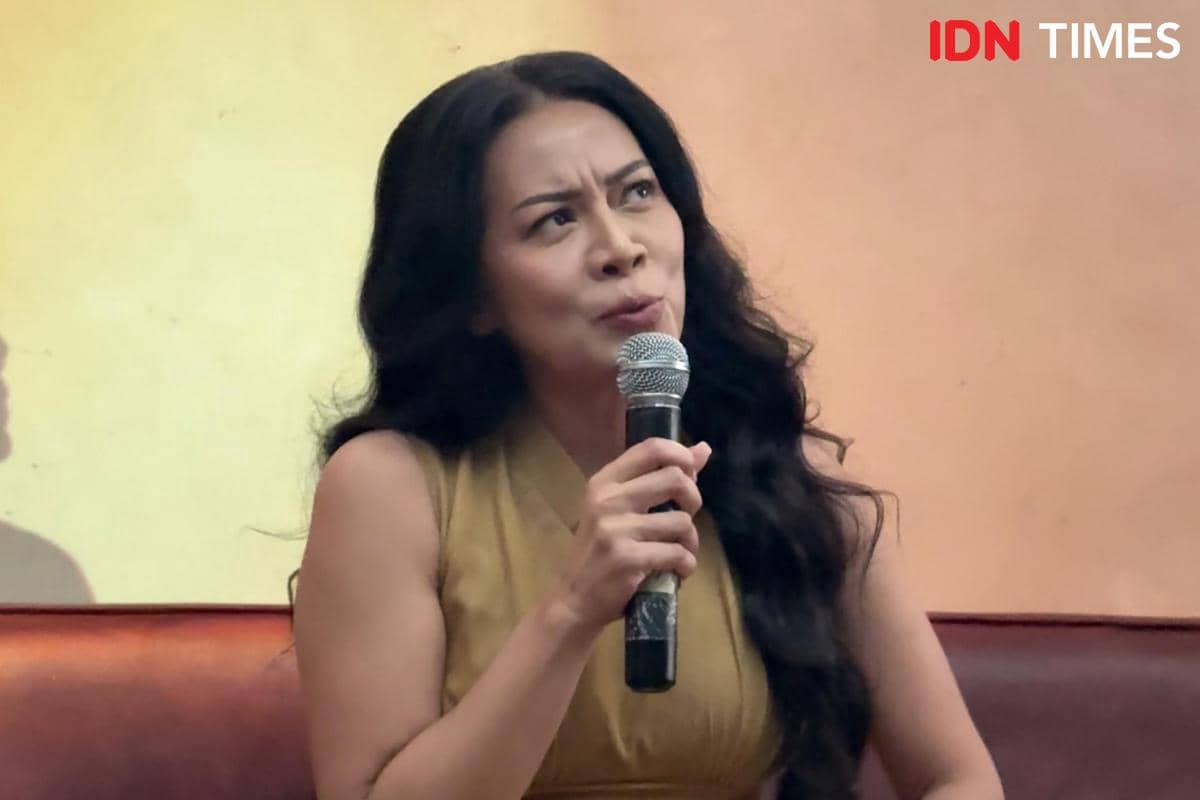 Seorang perempuan berambut panjang memegang mikrofon sambil berbicara dalam acara listening party album di ruangan dengan latar dinding krem.