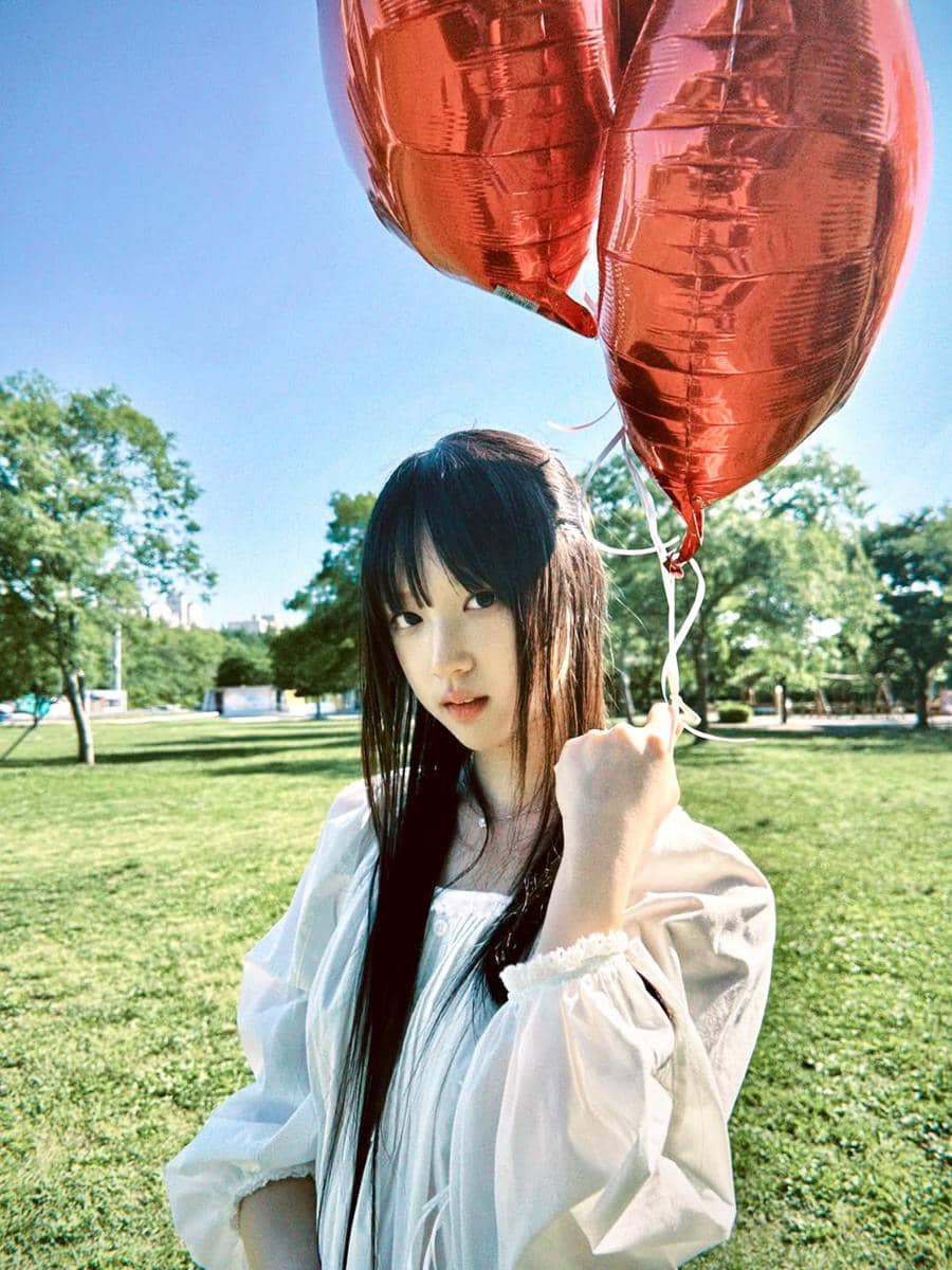 Seorang perempuan berambut panjang mengenakan blus putih memegang dua balon merah berbentuk hati di taman pada hari cerah.