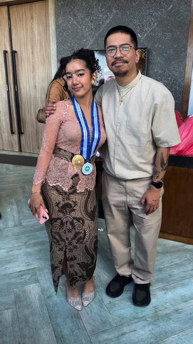 Seorang remaja mengenakan kebaya dan medali wisuda berpose bersama seorang pria berkacamata di acara kelulusan SMP.