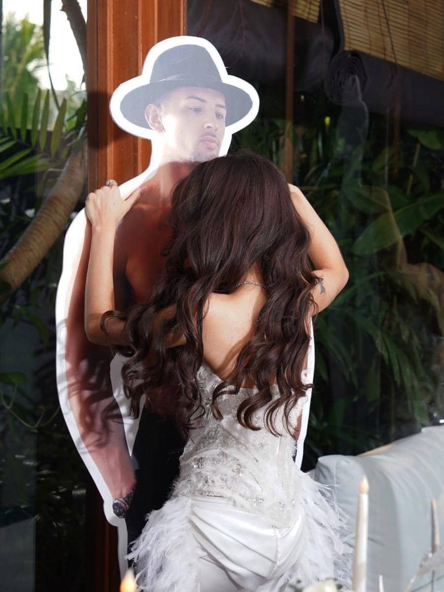 Seorang wanita bergaun putih dengan rambut panjang bergelombang memeluk papan bergambar pria bertopi hitam di acara bridal shower.