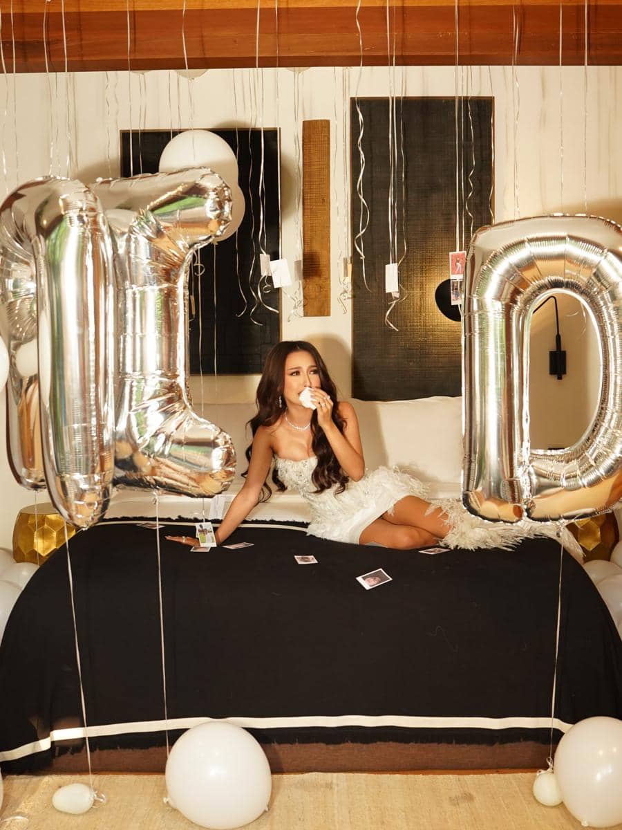 Seorang wanita mengenakan gaun putih duduk di atas tempat tidur dengan balon huruf perak besar dan dekorasi pesta bridal shower di sekitarnya.