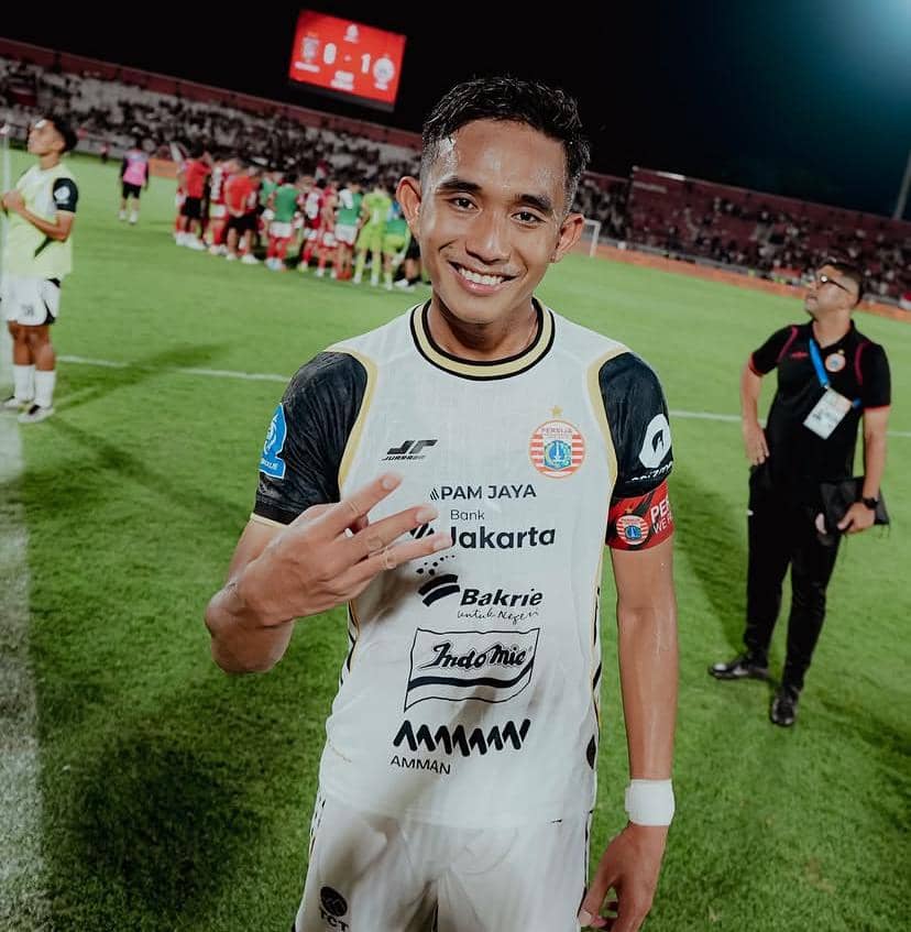 Seorang pemain sepak bola berseragam putih dengan logo Persija Jakarta tersenyum di lapangan setelah pertandingan malam hari.