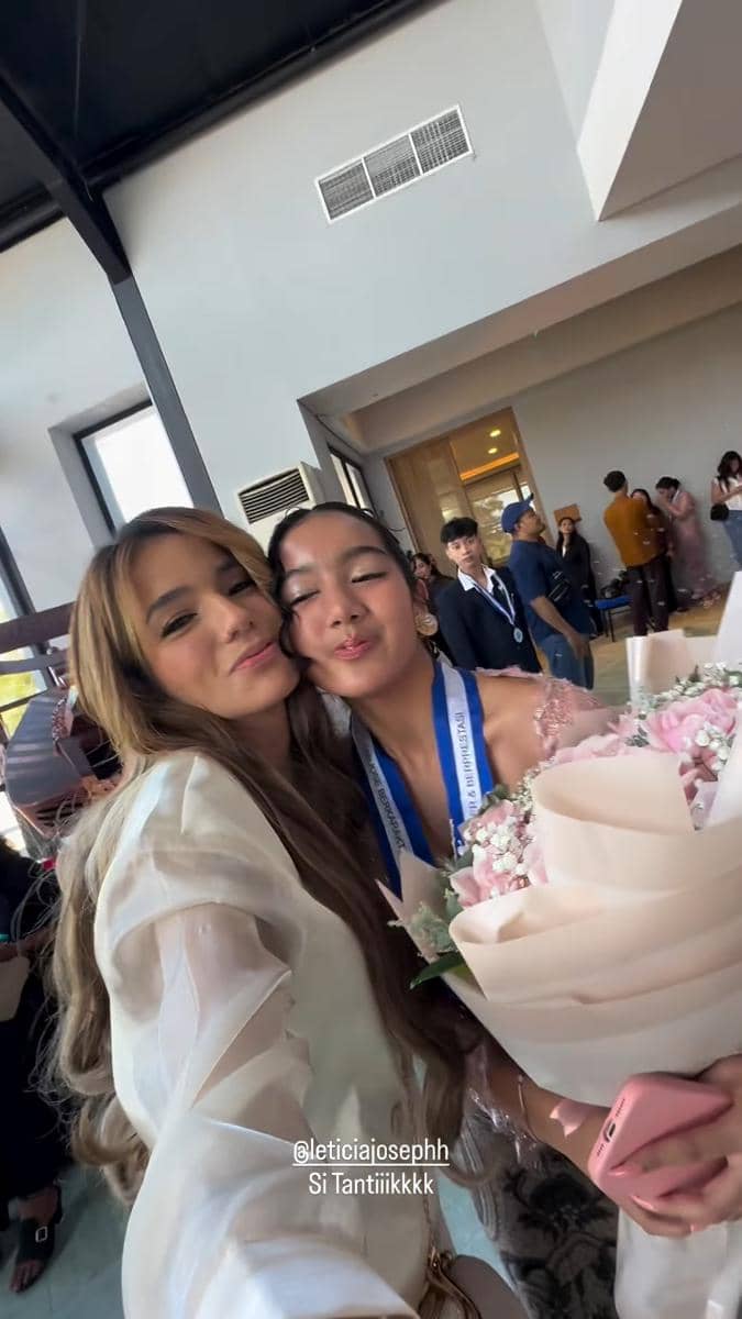 Seorang perempuan berpose bersama gadis muda yang mengenakan medali wisuda SMP dan memegang buket bunga berwarna merah muda.