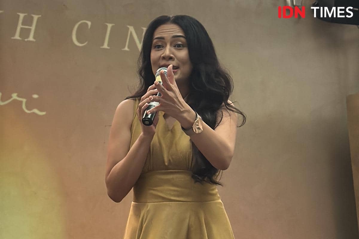 Seorang perempuan berambut panjang mengenakan gaun kuning sedang berbicara sambil memegang mikrofon di sebuah acara musik.