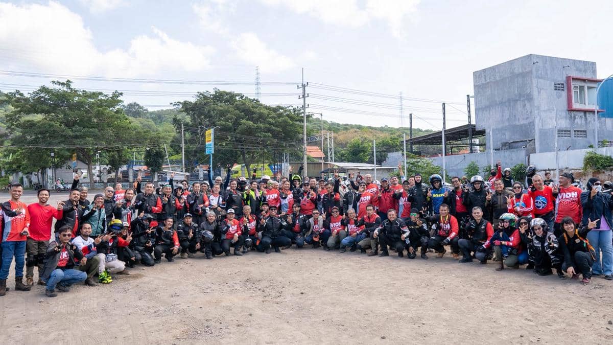 Ratusan pengendara motor berpose bersama di area terbuka dengan latar pepohonan dan bangunan di bawah langit cerah.