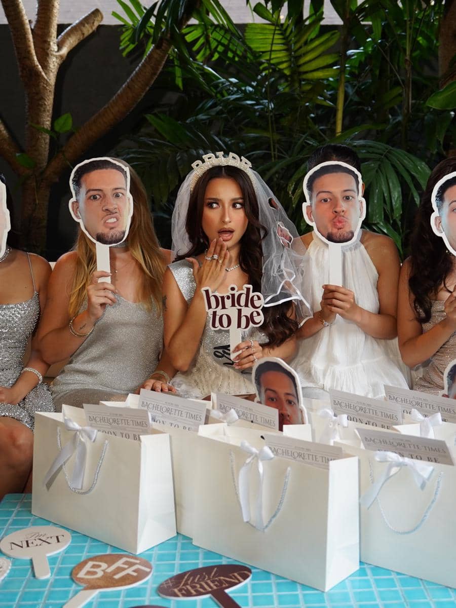 Seorang calon pengantin mengenakan veil dan mahkota bertuliskan 'bride to be' duduk bersama teman-temannya saat bridal shower dengan dekorasi lucu dan tas hadiah putih.