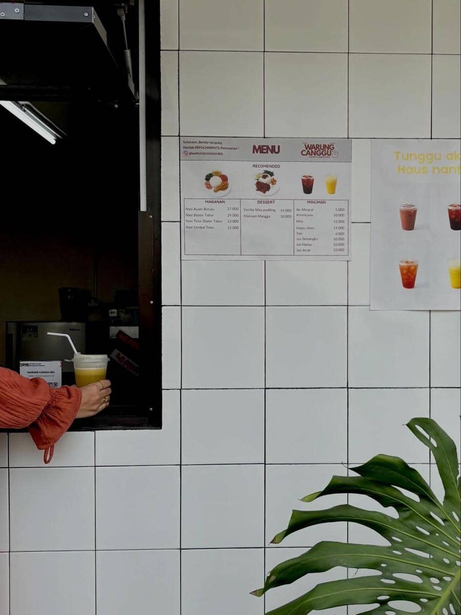 Tangan seseorang mengambil minuman dari jendela kecil di Warung Canggu dengan latar dinding ubin putih dan menu yang tertempel.