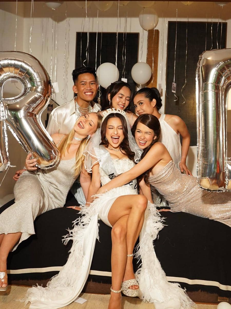 Sekelompok teman berpakaian elegan berpose ceria di pesta bridal shower dengan balon perak dan dekorasi balon putih di latar belakang.