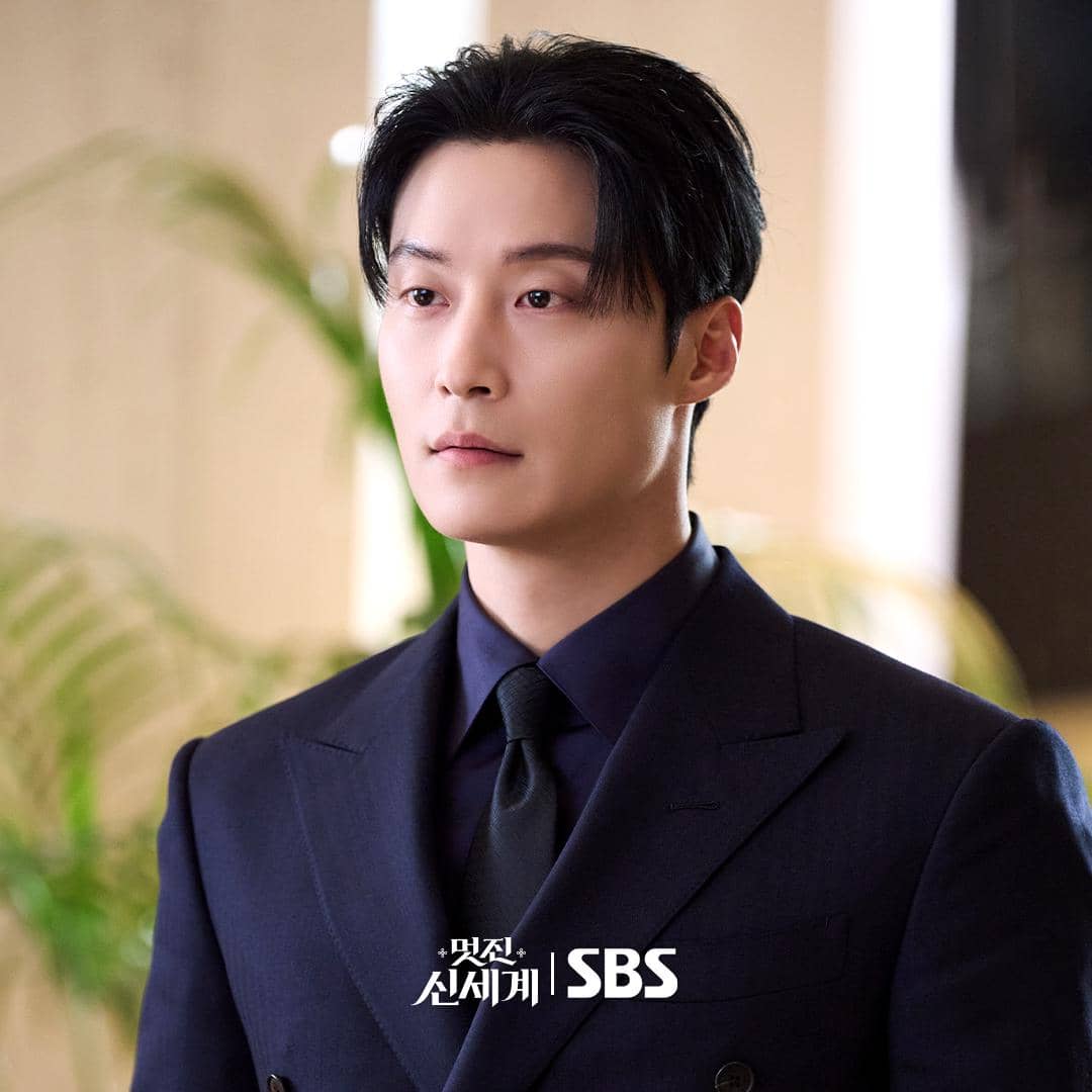 Seorang pria berpenampilan rapi mengenakan setelan jas hitam dan dasi gelap dalam cuplikan drama My Royal Nemesis produksi SBS.
