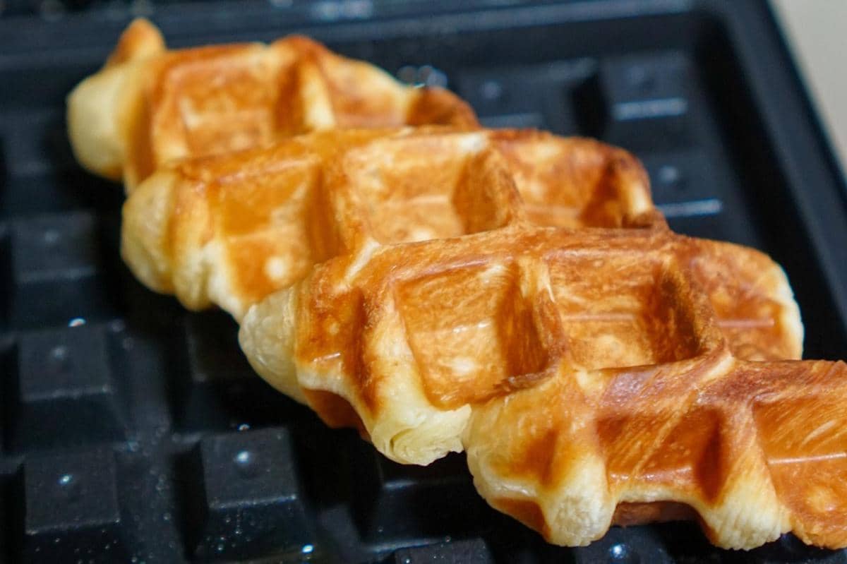 Beberapa croffle berwarna keemasan sedang dipanggang di atas alat pemanggang waffle dengan tekstur renyah di permukaannya.