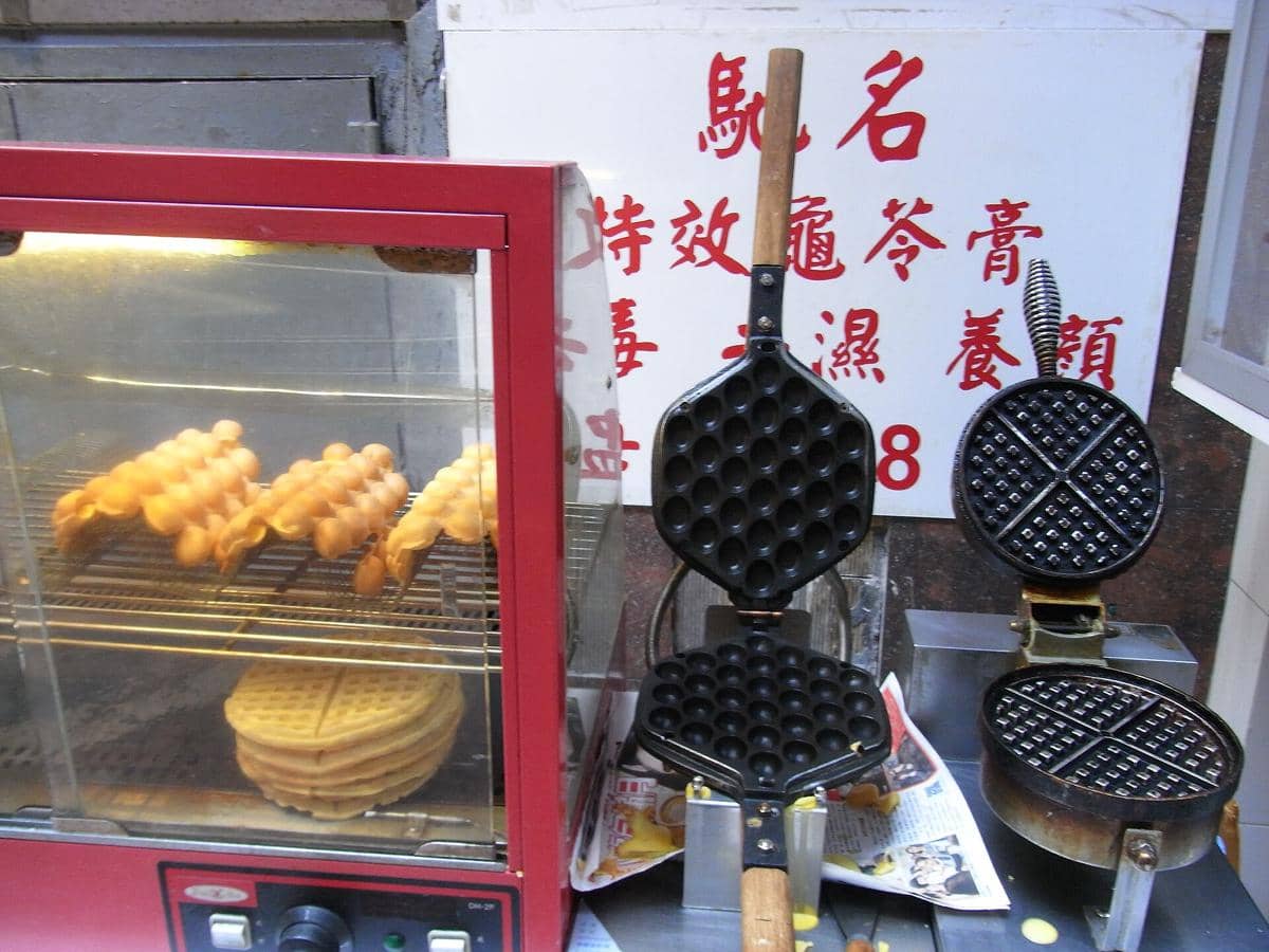 Cetakan waffle berbentuk bulat dan heksagonal dengan adonan waffle yang sedang dipanggang di etalase kaca berwarna merah.