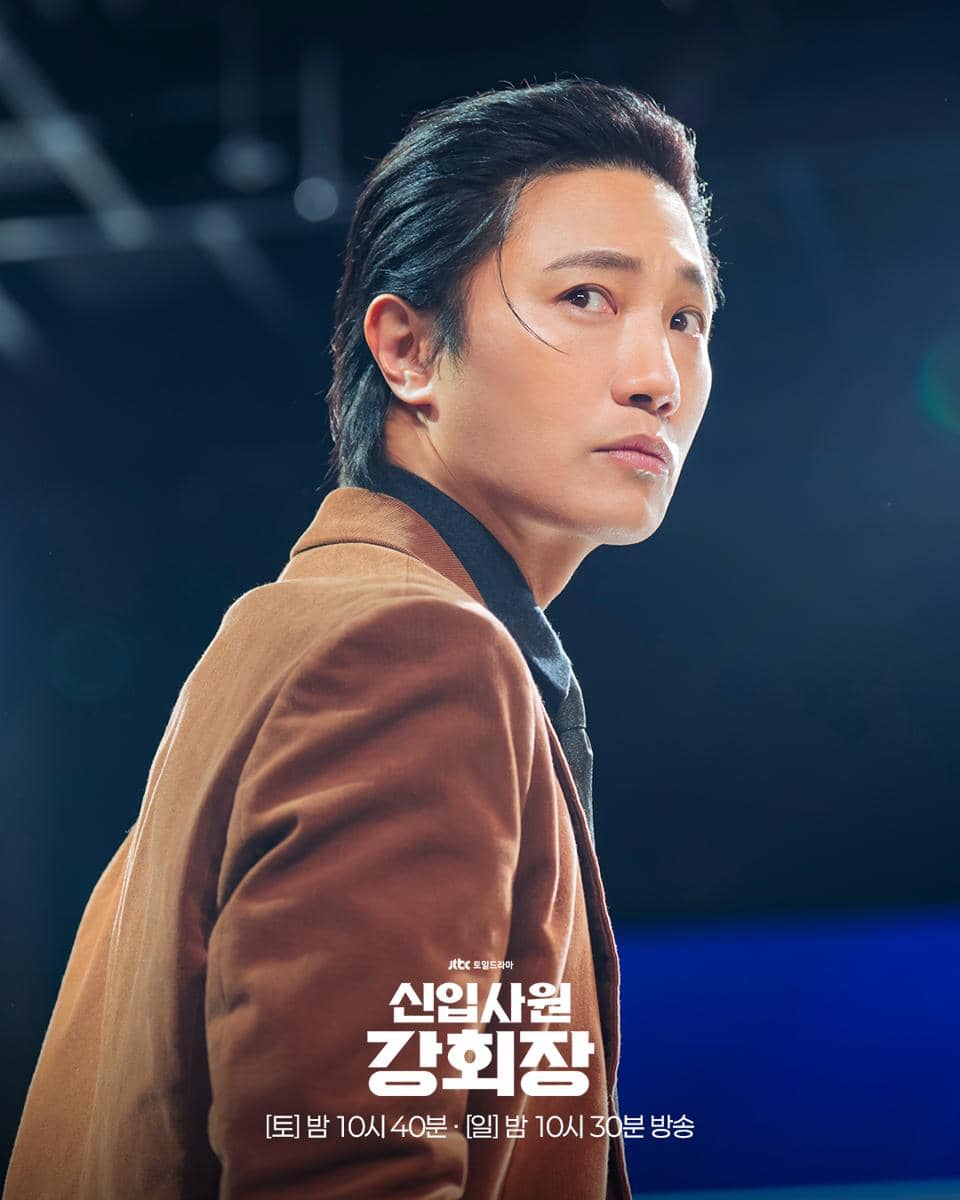 Seorang aktor mengenakan jas cokelat dan kemeja hitam sedang menatap ke samping dalam cuplikan drama Reborn Rookie.