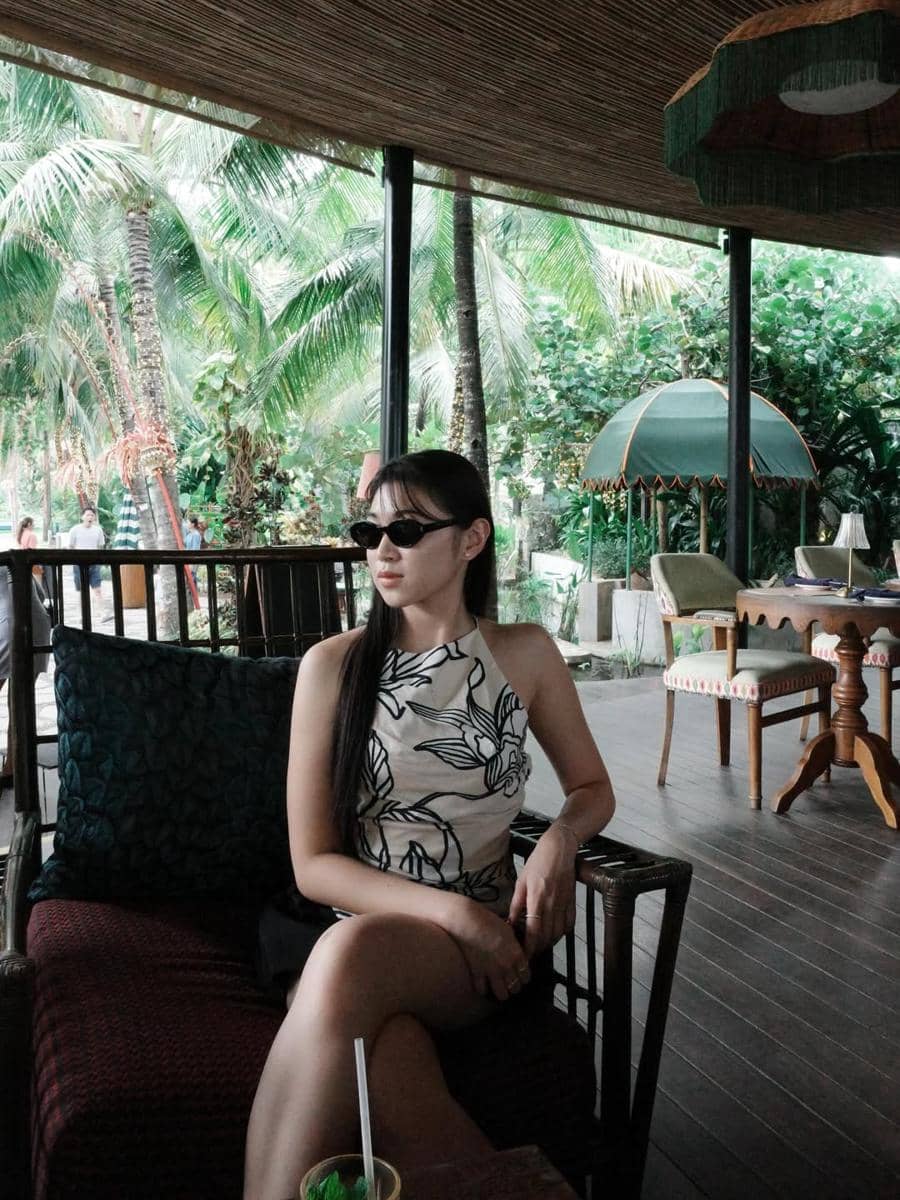 Seorang wanita mengenakan atasan tanpa lengan bermotif duduk santai di kafe bernuansa tropis dengan latar pepohonan kelapa.