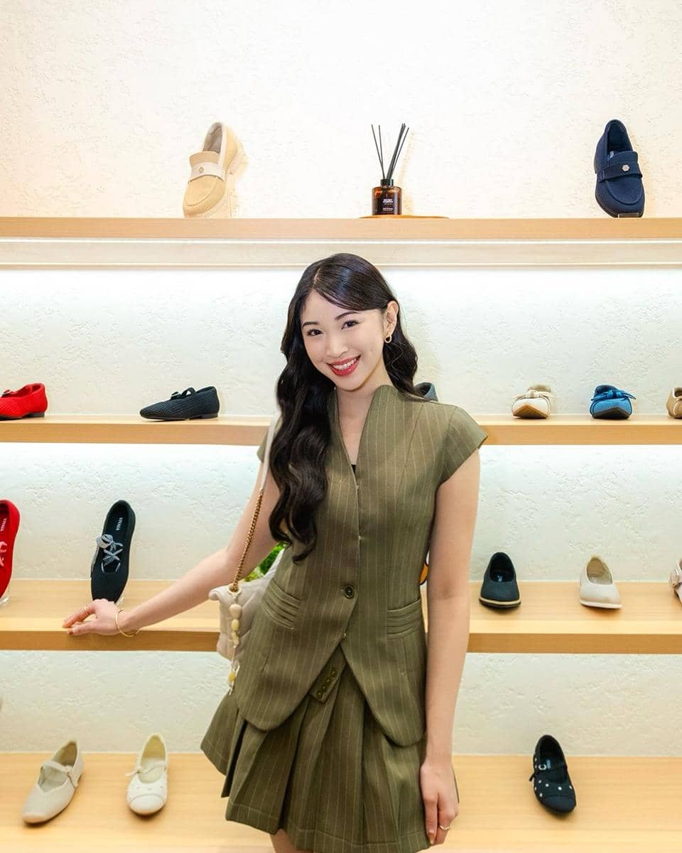 Seorang wanita berambut panjang mengenakan setelan hijau bergaris berdiri di depan rak pajangan sepatu berbagai warna di dalam toko.