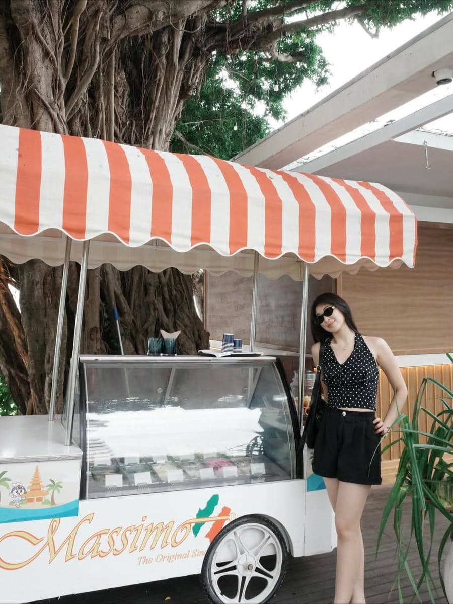 Seorang wanita bergaya kasual mengenakan atasan hitam bermotif polkadot dan celana pendek hitam berdiri di samping gerobak es krim Massimo.