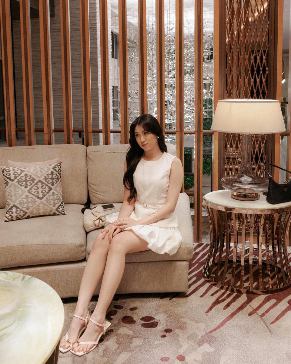 Seorang wanita mengenakan gaun putih duduk di sofa ruang tamu modern dengan pencahayaan hangat dan dekorasi elegan di sekitarnya.