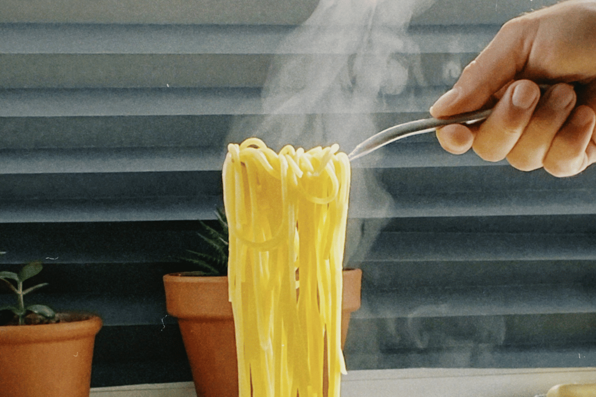Tangan memegang garpu dengan mie ramen panas yang mengepul di depan jendela dengan pot tanaman di latar belakang.