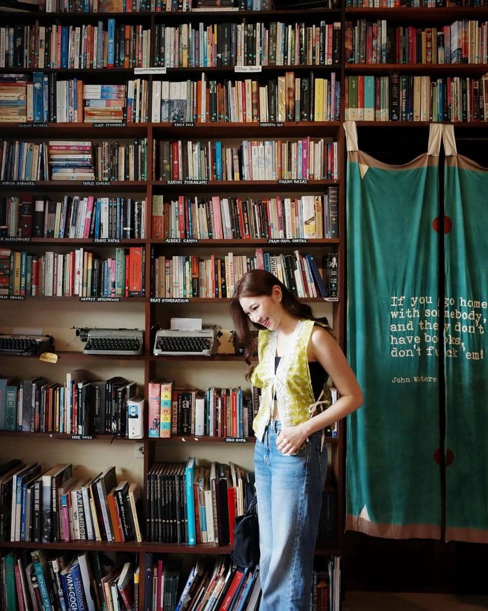 Seorang wanita bergaya kasual dengan rompi kuning dan jeans berdiri di depan rak buku besar di dalam ruangan yang penuh buku.