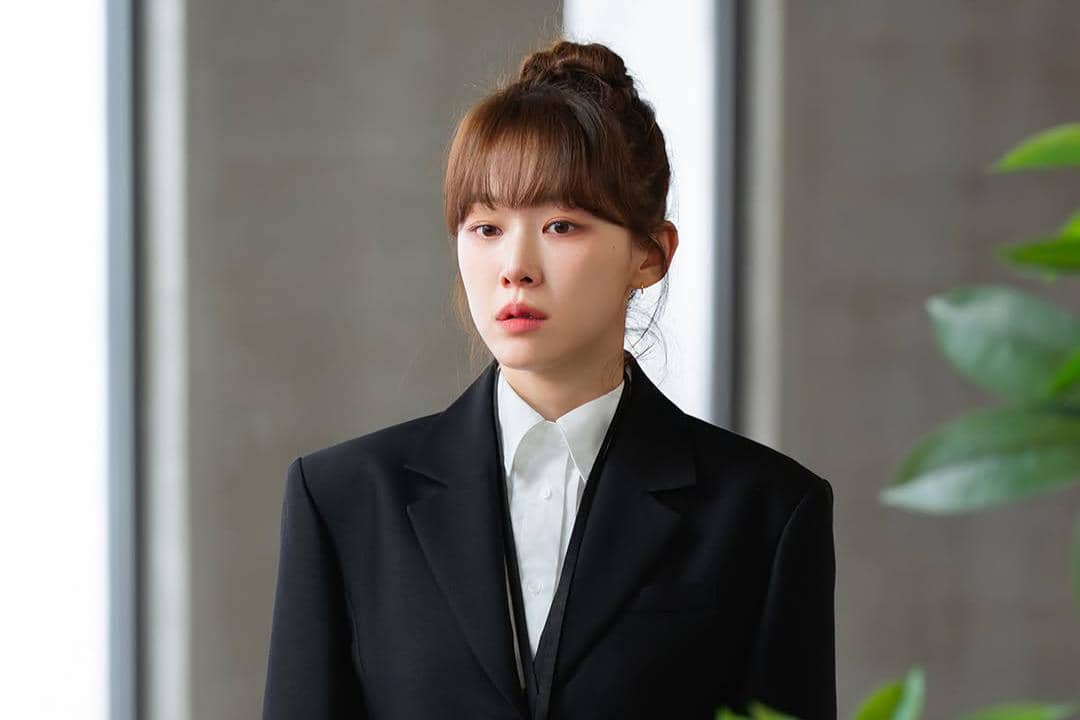 Seorang wanita berambut cokelat dengan poni mengenakan setelan jas hitam dan kemeja putih berdiri di dalam ruangan modern.