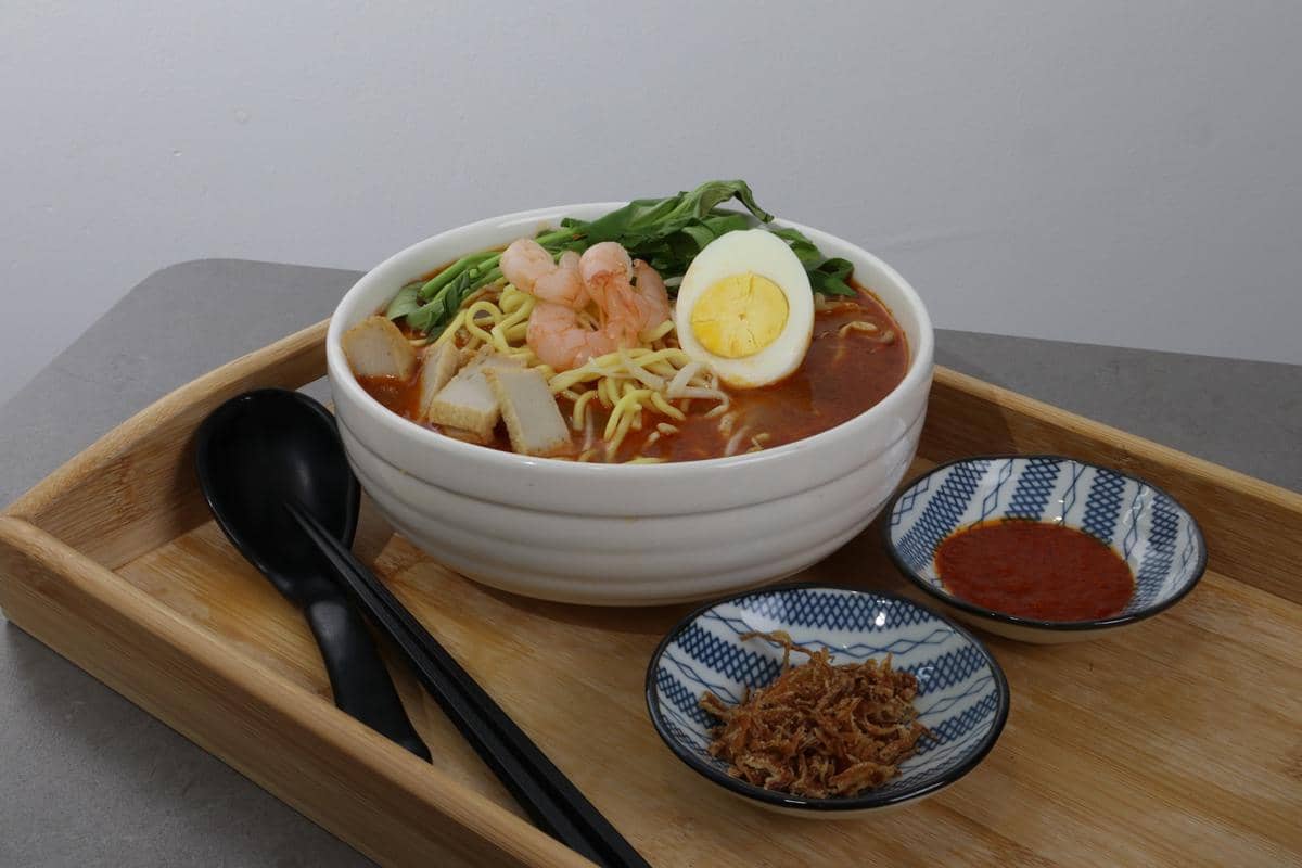 Semangkuk ramen kari merah dengan udang, telur rebus, sayuran, dan potongan fish cake disajikan di atas nampan kayu dengan sambal dan bawang goreng.