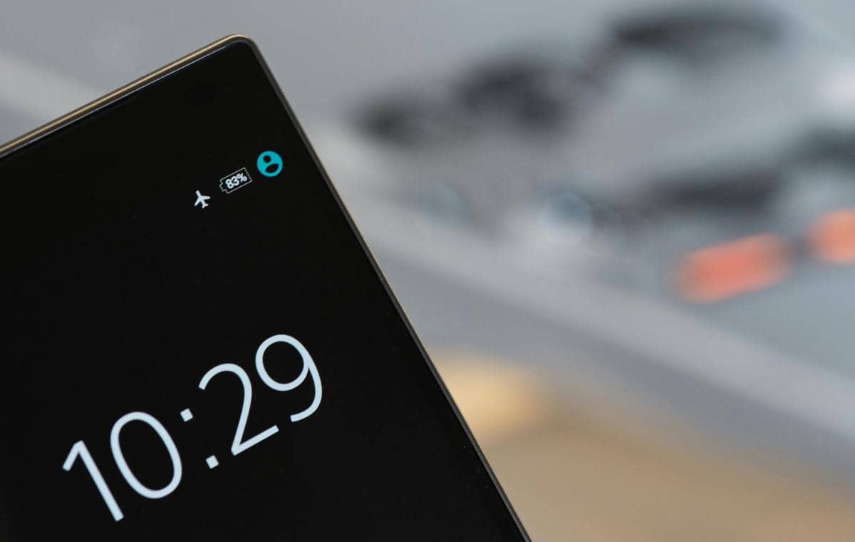 Bagian sudut atas layar smartphone hitam yang menampilkan jam digital 10:29, persentase baterai 83%, ikon profil biru, dan indikator airplane mode (mode pesawat) aktif, dengan latar belakang papan ketik yang kabur.