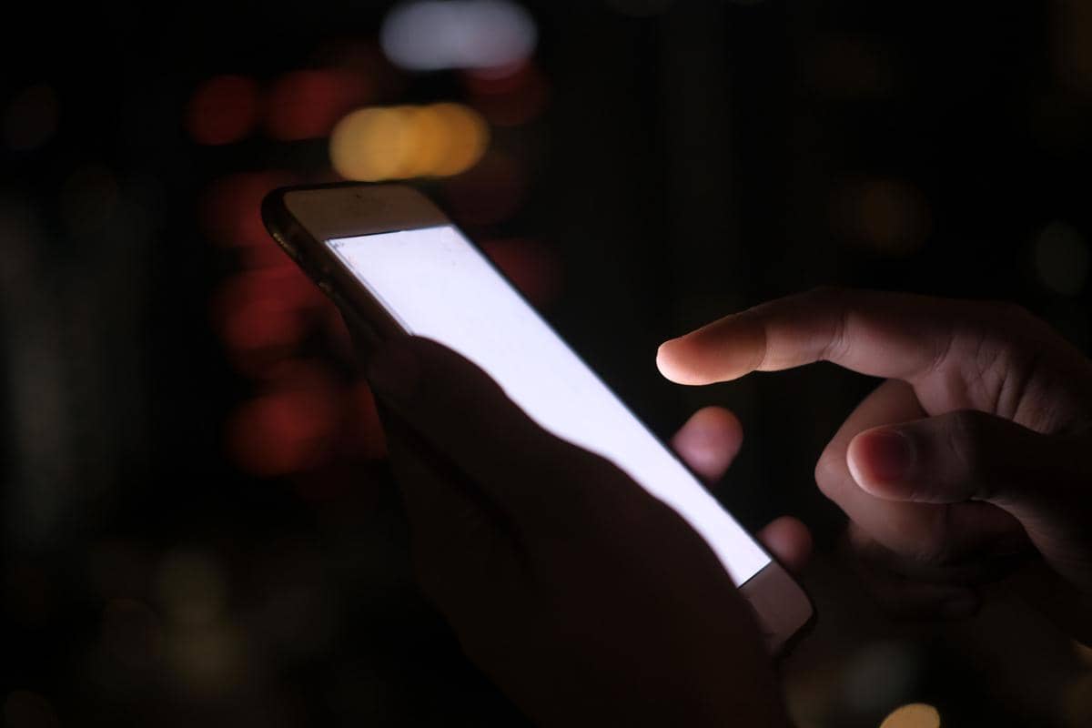 Jari telunjuk seseorang bersiap menyentuh layar smartphone yang menyala terang putih di malam hari, dengan latar belakang lampu kota yang buram (bokeh) berwarna kemerahan dan kekuningan.