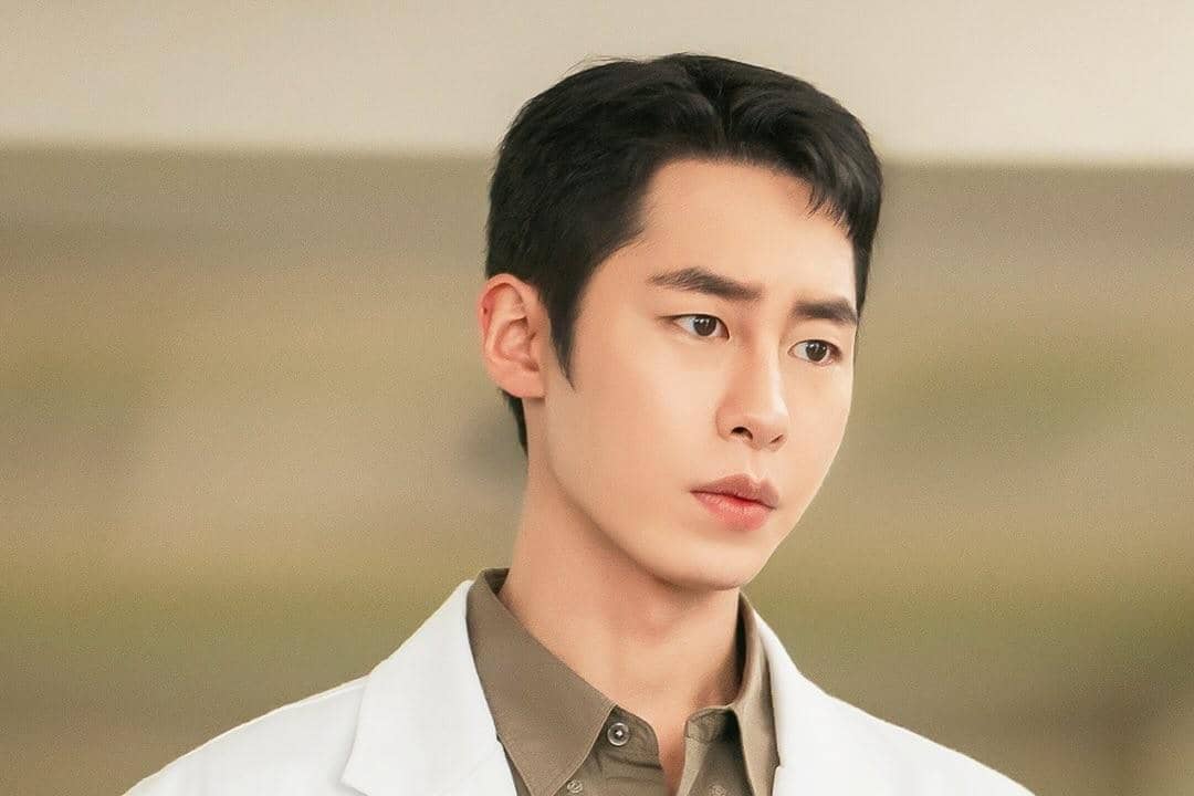 Seorang pria mengenakan jas dokter putih dan kemeja cokelat muda tampak serius dalam cuplikan drama Doctor On The Edge.