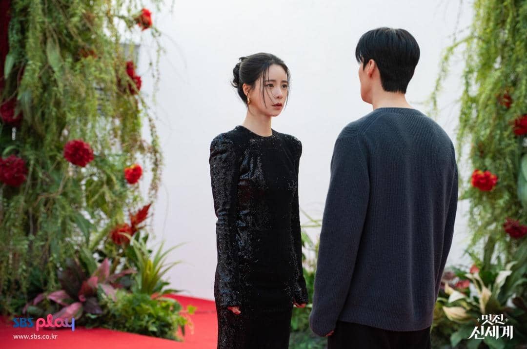 Dua karakter dalam drama My Royal Nemesis berdiri saling berhadapan di taman dengan latar bunga merah dan tanaman hijau.