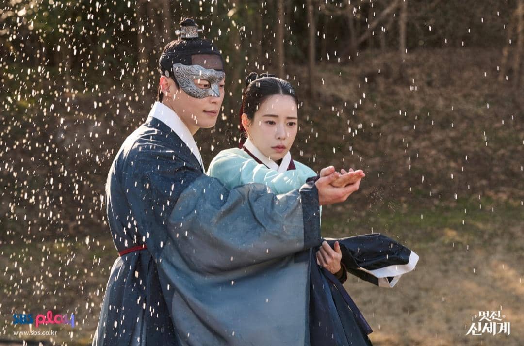 Dua karakter mengenakan hanbok tradisional Korea berdiri di bawah percikan air, salah satunya memakai topeng perak dan memegang tangan pasangannya.
