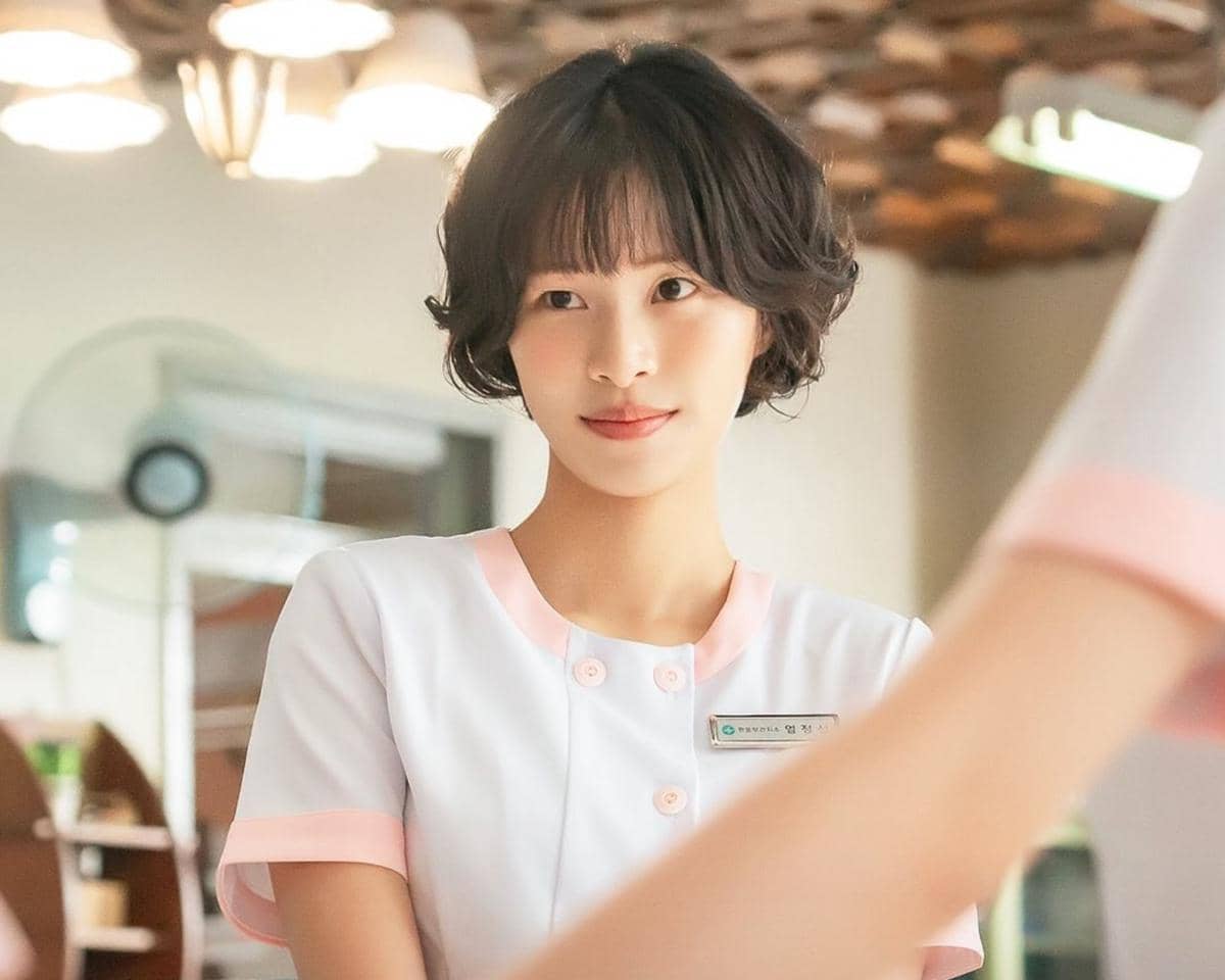 Seorang perempuan berambut pendek mengenakan seragam perawat putih dengan aksen merah muda sedang berada di dalam ruangan terang.