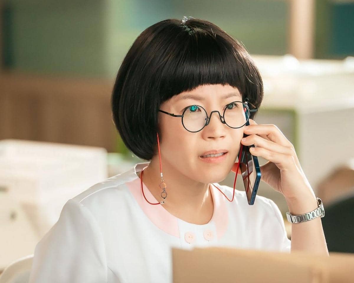 Seorang wanita berambut pendek mengenakan kacamata bulat sedang berbicara di telepon di sebuah ruangan dengan pencahayaan hangat.