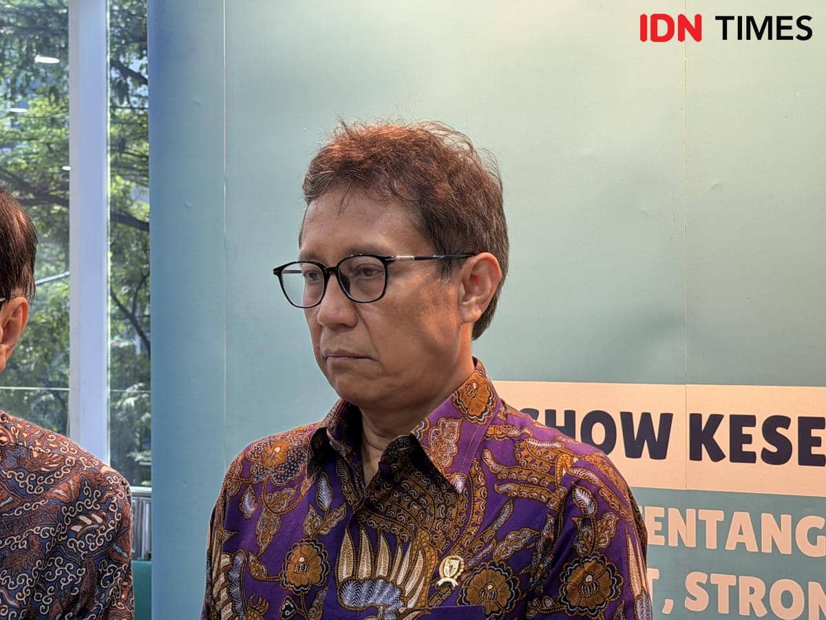 Seorang pria mengenakan batik ungu bermotif cokelat dan kacamata berdiri di depan latar bertuliskan IDN Times dan teks bertema kesehatan.