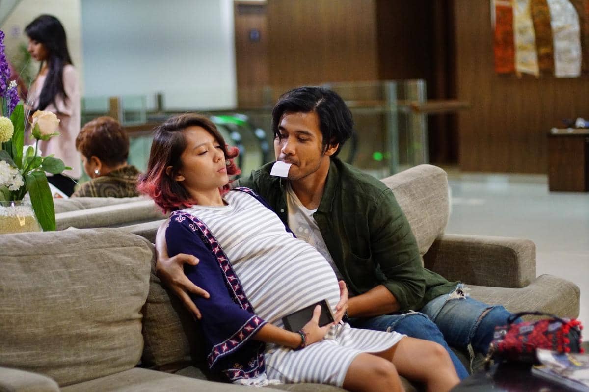 Seorang wanita hamil duduk di sofa bersama seorang pria yang menemaninya di ruang tunggu, keduanya tampak berinteraksi dengan ekspresi serius.