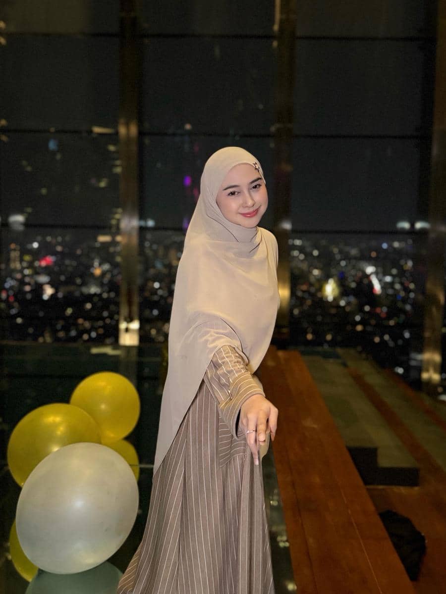 Seorang wanita berhijab berpose sambil tersenyum di malam hari dengan latar belakang pemandangan kota dan balon berwarna kuning serta putih.
