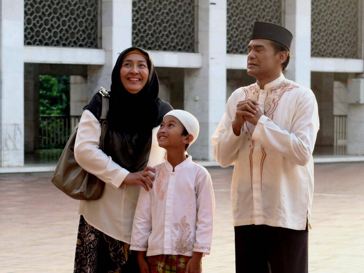 Seorang pria, wanita, dan anak laki-laki mengenakan busana muslim berdiri di halaman masjid dengan ekspresi hangat dan khusyuk.