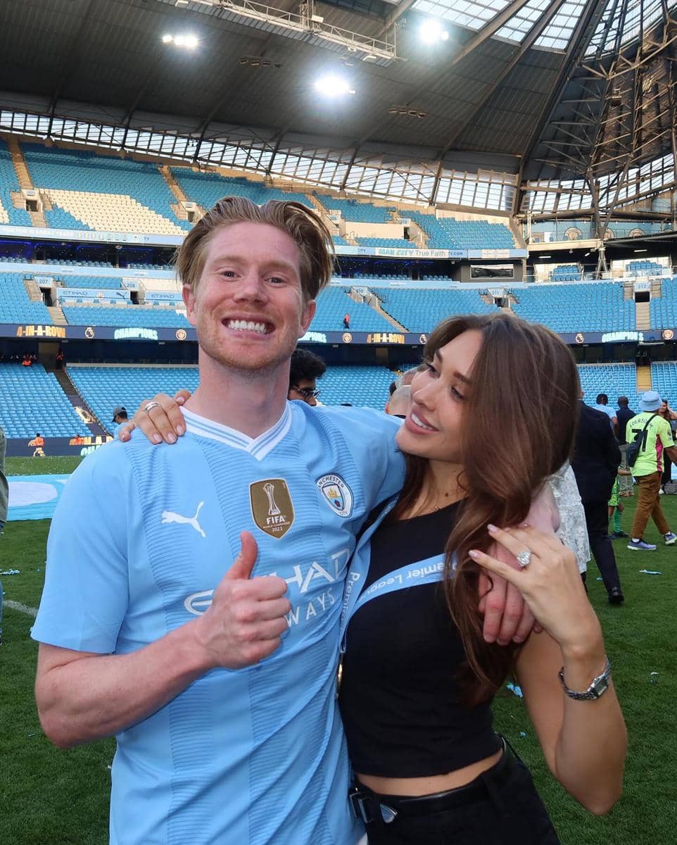 Seorang pemain sepak bola mengenakan jersey biru muda berpose dengan seorang wanita di stadion setelah pertandingan.