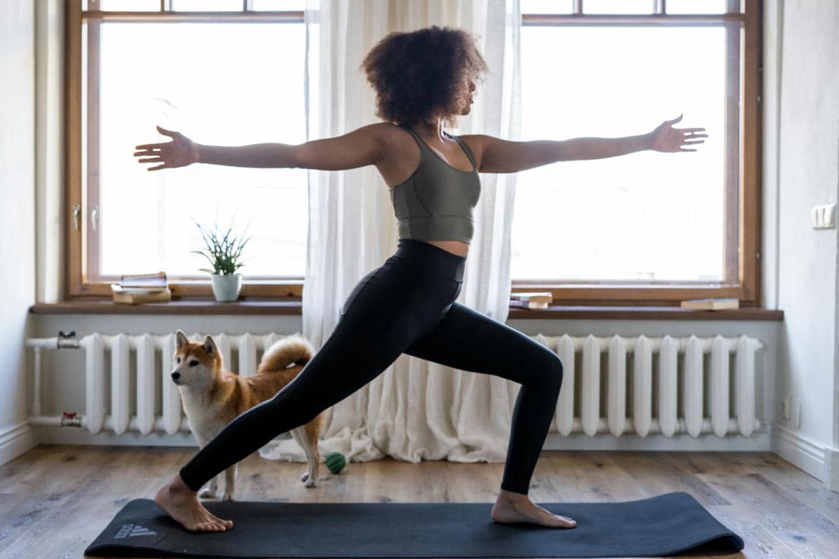 Seorang perempuan melakukan pose yoga di atas matras hitam di ruang tamu dengan seekor anjing berdiri di dekatnya.