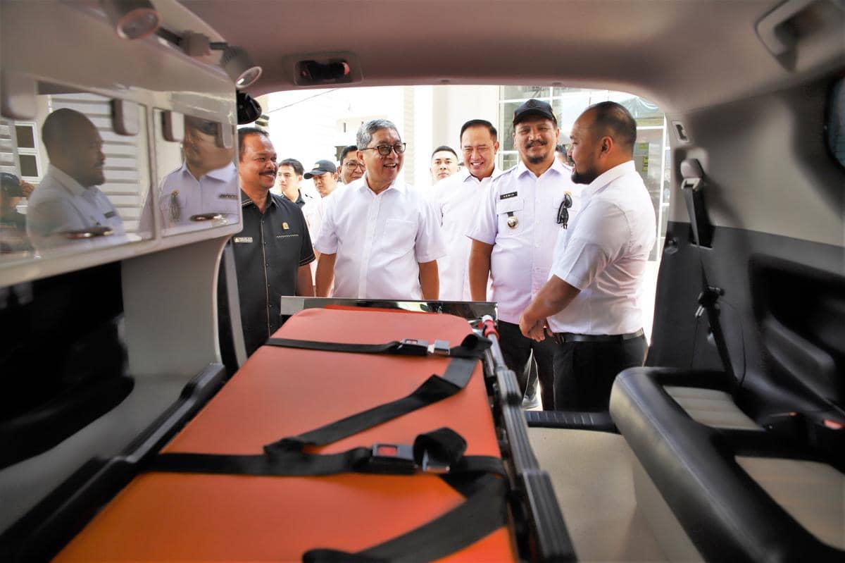 Beberapa pria berpakaian dinas putih dan hitam berdiri di dekat bagian belakang mobil ambulans yang berisi tandu oranye.