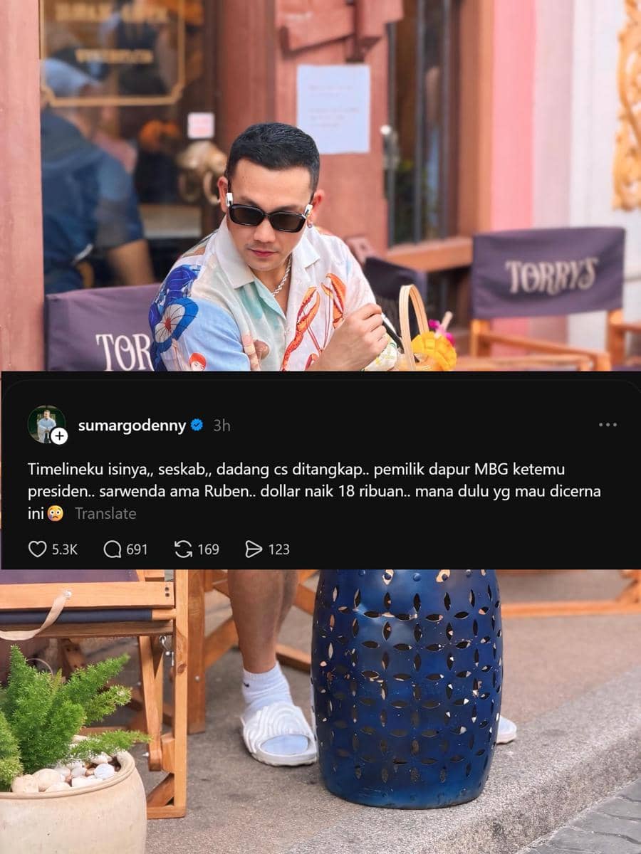 Seorang pria mengenakan kemeja bermotif warna-warni dan kacamata hitam duduk di kafe outdoor dengan minuman di meja di depannya.