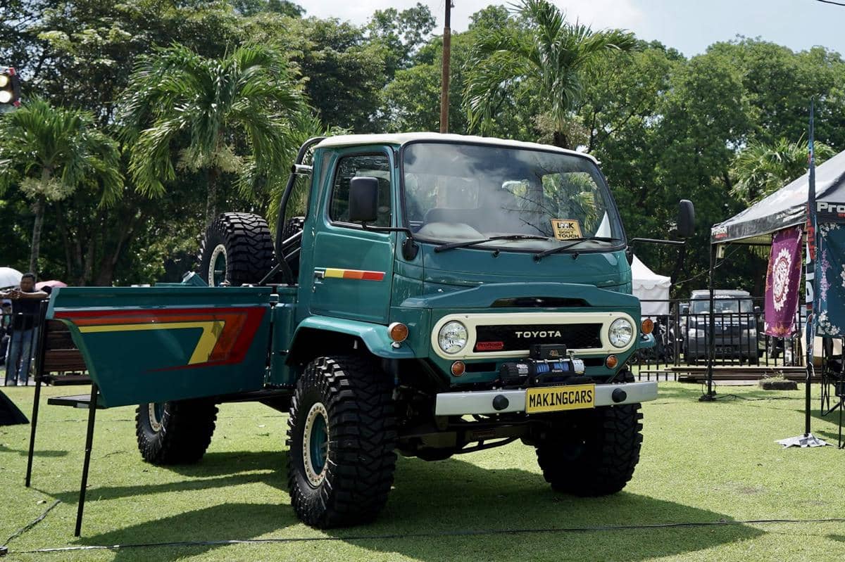 Mobil Toyota klasik hasil modifikasi berwarna hijau tua dengan bak terbuka dipamerkan di area luar ruangan dengan latar pepohonan.