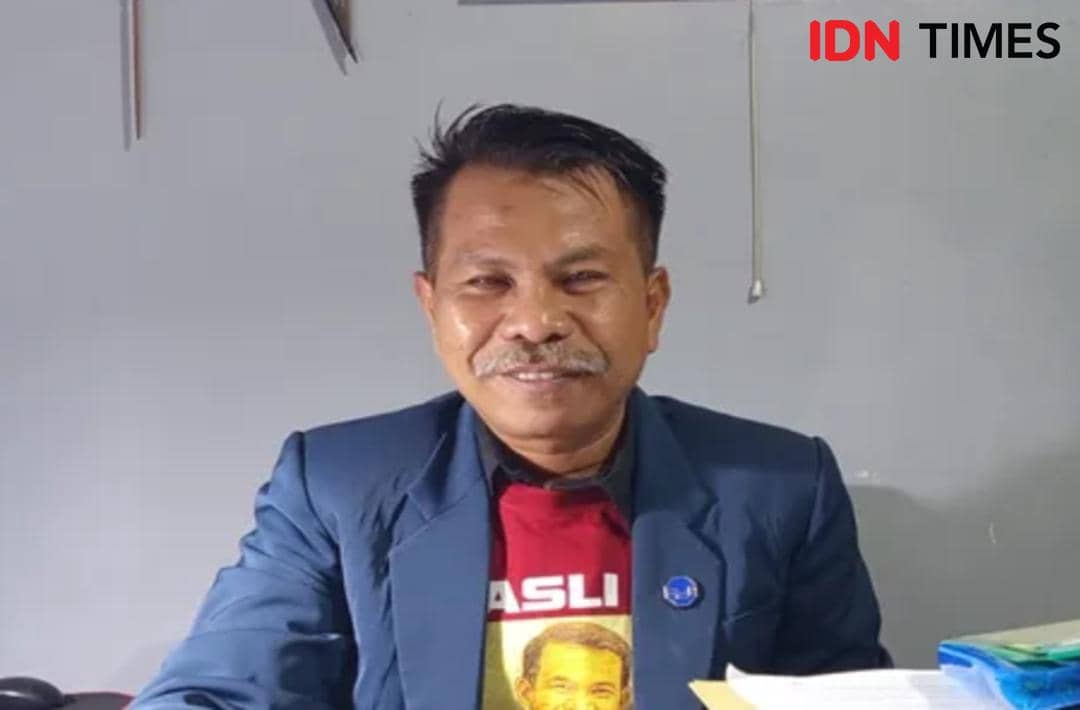 Seorang pria mengenakan jas biru dan kaus merah sedang duduk di meja dengan tumpukan berkas di depannya di sebuah ruangan.