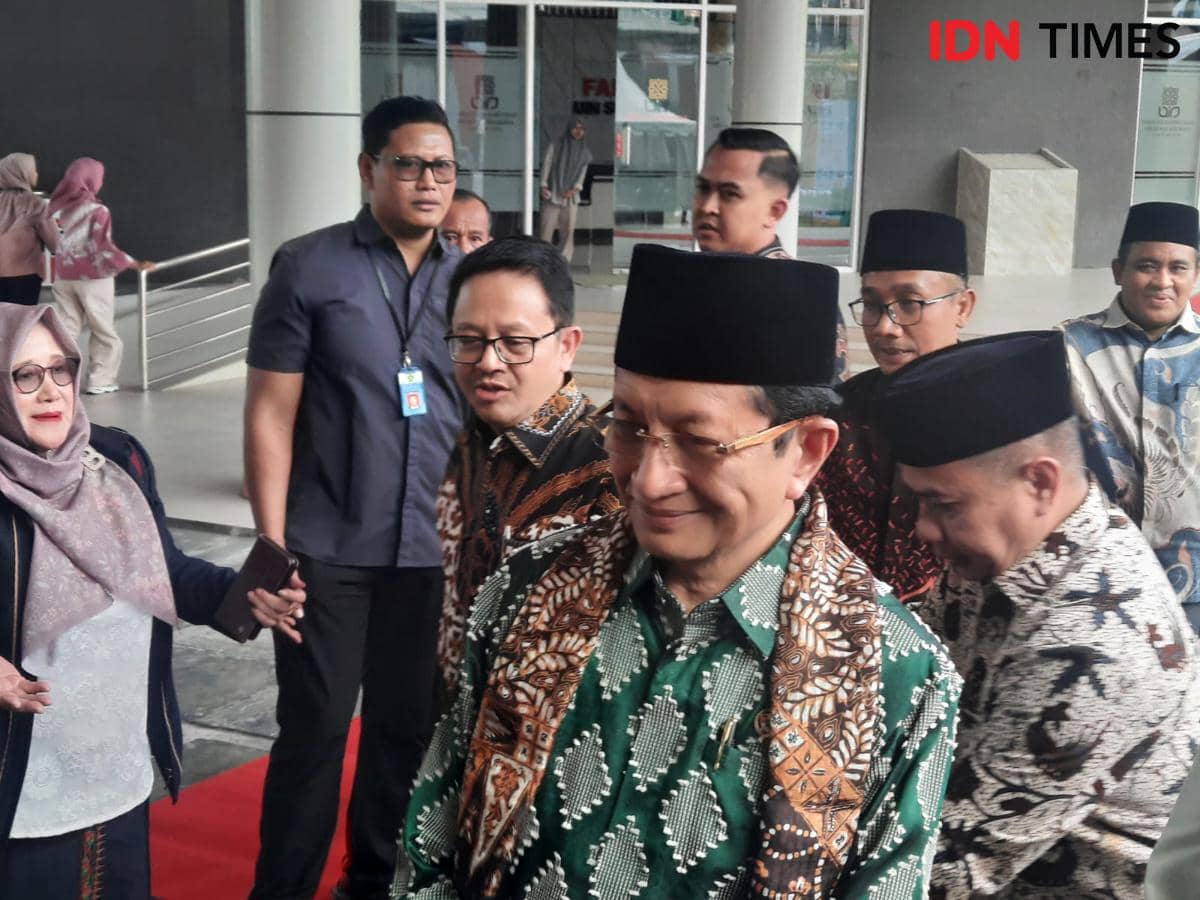 Seorang pejabat mengenakan batik hijau dan peci hitam berjalan di karpet merah, dikelilingi beberapa orang di area gedung modern.