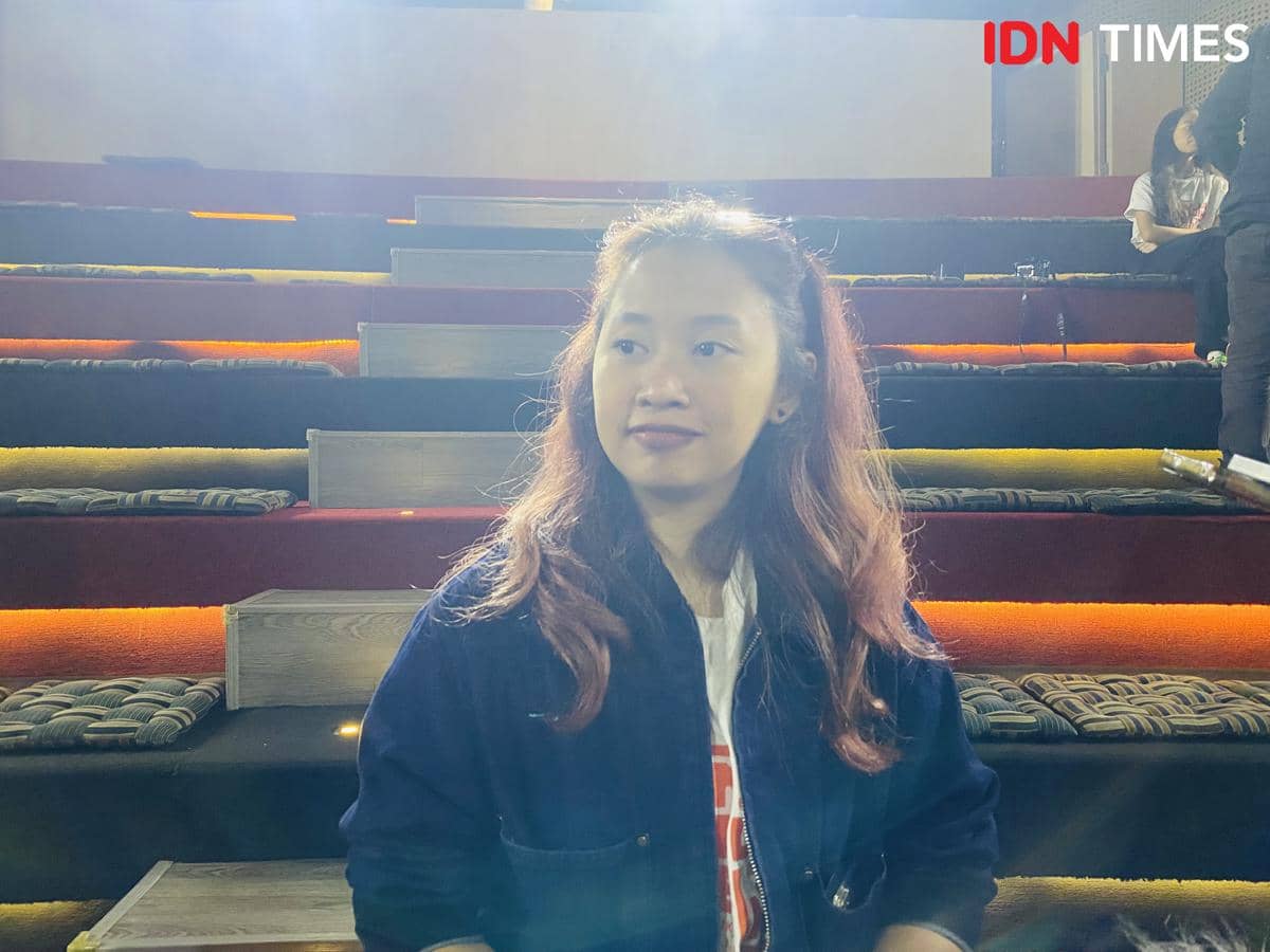 Seorang perempuan berambut panjang mengenakan jaket biru duduk di area tribun dengan pencahayaan hangat di sekitarnya.