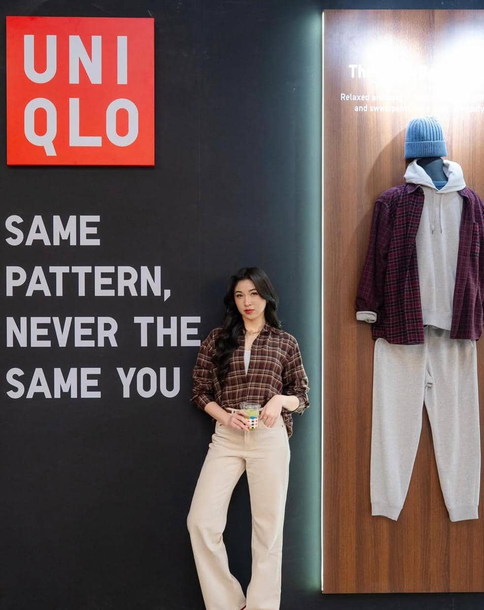 Seorang wanita berpose di depan dinding bertuliskan slogan Uniqlo dengan outfit kasual kemeja kotak-kotak dan celana krem.