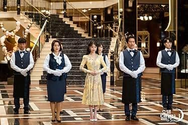 Para pemeran drama Hotel del Luna berdiri di lobi hotel mewah dengan tangga besar di latar belakang, mengenakan seragam elegan.