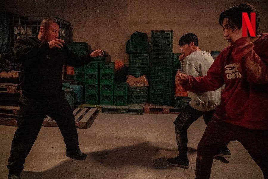 Tiga pria bersiap bertarung di ruangan gelap dengan tumpukan peti plastik di belakang mereka dalam adegan drama Bloodhounds.