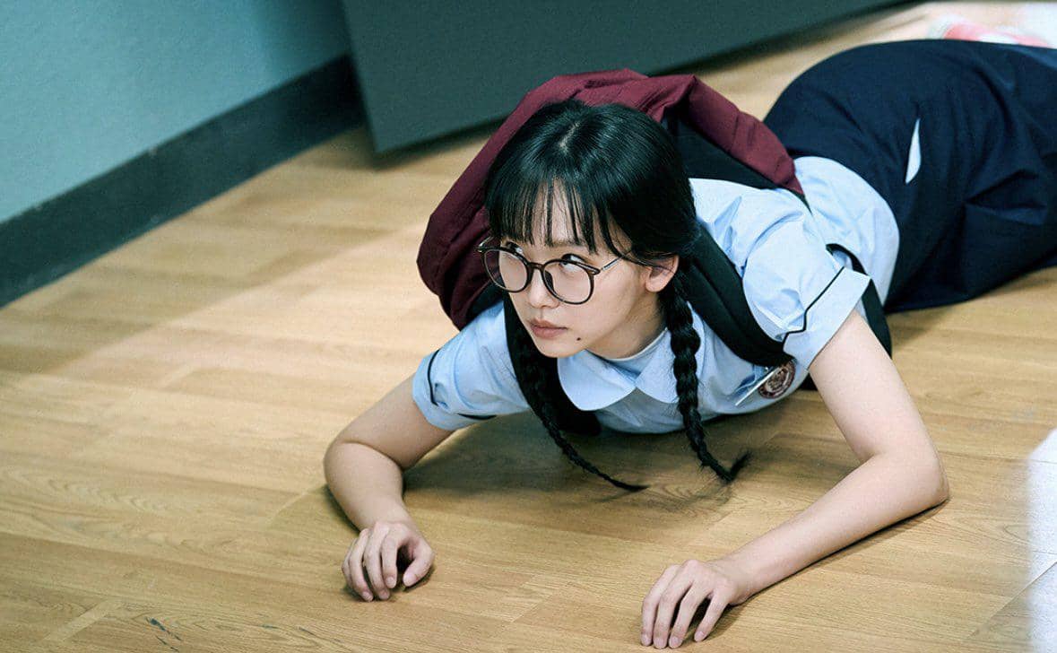 Seorang siswi berkacamata dengan seragam sekolah dan ransel merah merangkak di lantai kayu dalam adegan drama Teach You a Lesson.